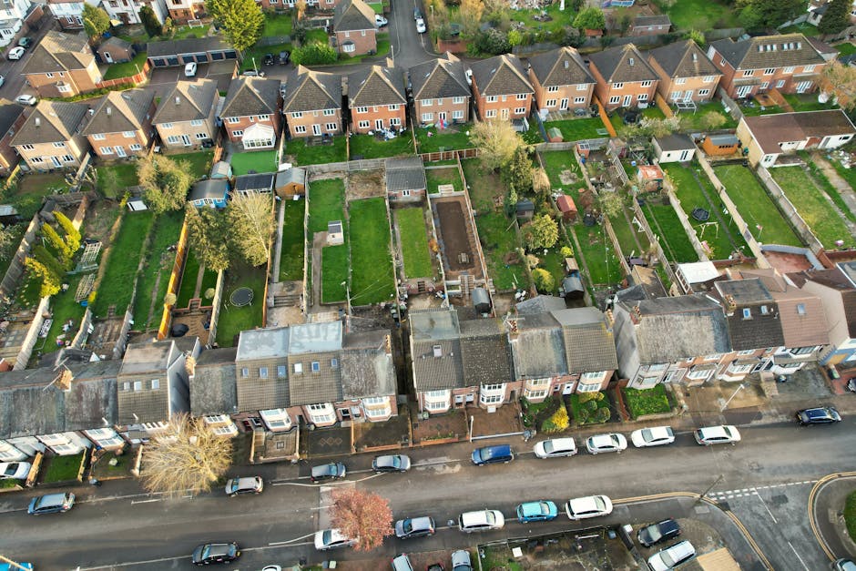 An aerial view of a suburban residential area showing a row of terraced houses with front gardens facing the street. Each garden features well-maintained lawns, bordered by wooden or brick fences, wit